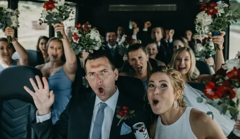 Bride, groom, and wedding party celebrating with bouquets raised inside a charter bus