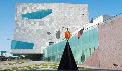 Exterior of a modern museum building with a textured metallic facade and a large colorful mobile sculpture in the foreground