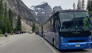 Van Hool motorcoach parked on a mountain road with dramatic rocky peaks and a group of passengers nearby