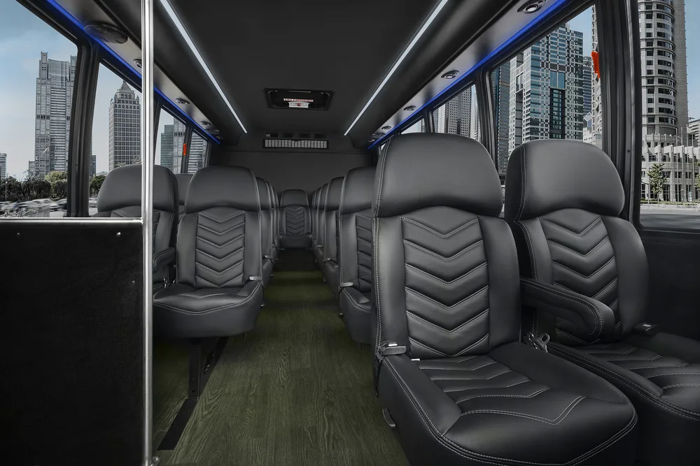Interior of a charter bus with rows of black leather seats, wood-grain flooring, and LED lighting along the ceiling