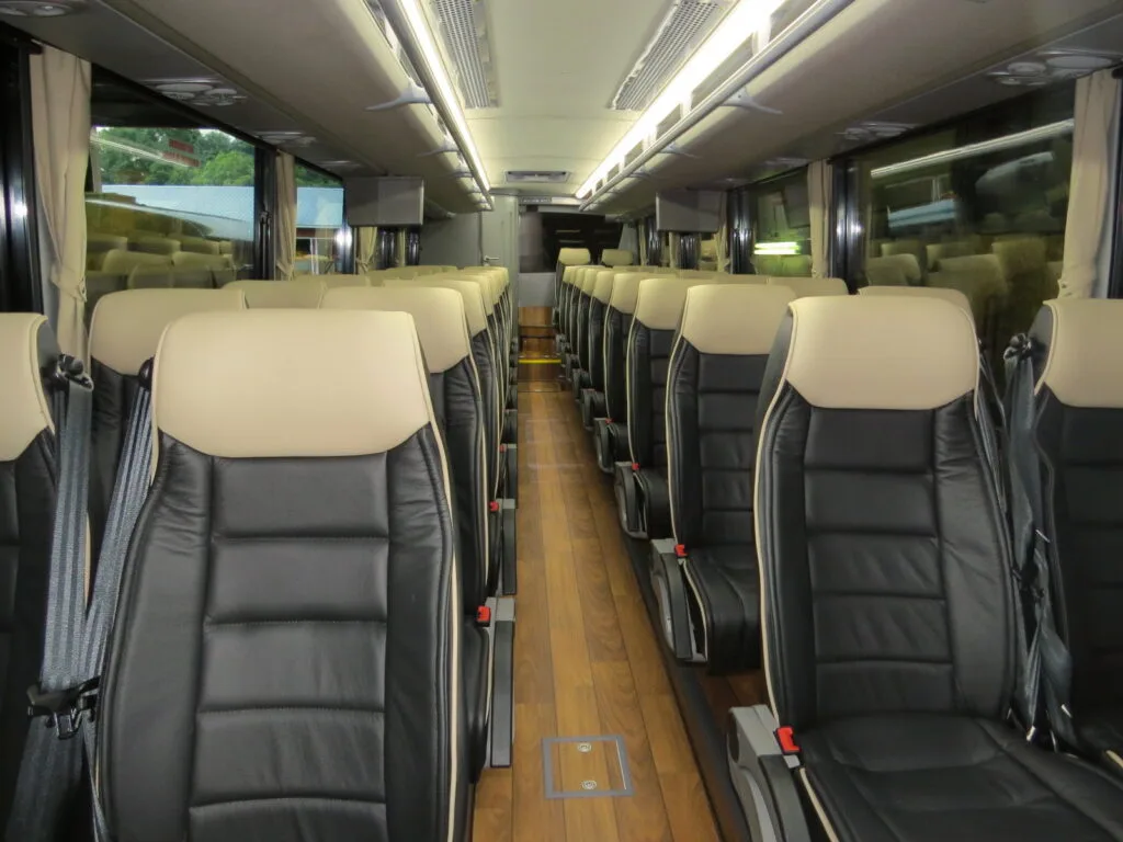 Interior of a charter bus cabin with rows of leather seats with seatbelts, overhead lighting, and a wood-look floor aisle