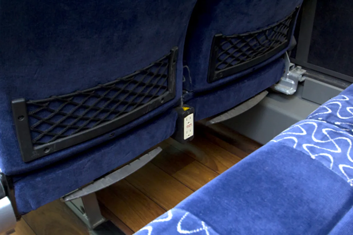 Blue upholstered charter bus seats with seat-back net pockets and an electrical outlet mounted between rows