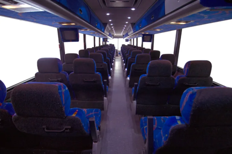 Interior of a motorcoach with rows of padded blue and gray seats, overhead lighting, and onboard monitors lining the aisle