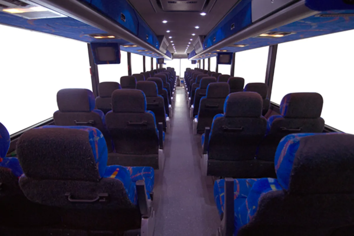 Interior of a motorcoach with rows of padded blue and gray seats, overhead lighting, and onboard monitors lining the aisle