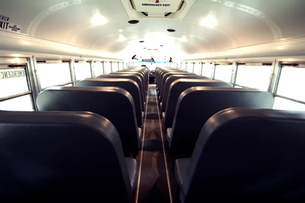 Interior view of a school bus showing rows of dark seats, a center aisle, and an emergency exit at the rear