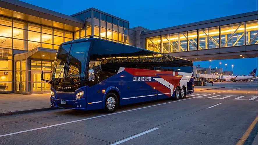 Lorenz Bus Service Prevost motorcoach parked outside an airport terminal at dusk, with aircraft visible in the background