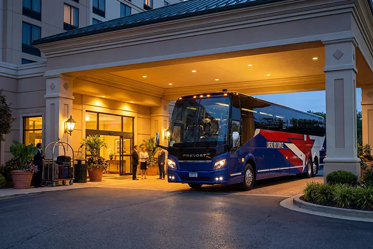Lorenz Bus Service Prevost motorcoach parked under a hotel porte-cochère at dusk, with passengers and a bellhop nearby