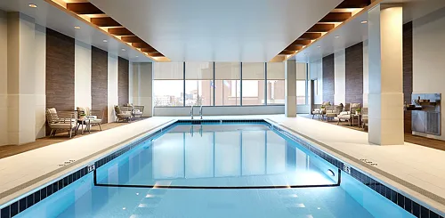 Indoor hotel pool with calm blue water, surrounded by lounge chairs and large windows with a city view