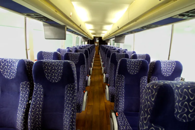 Interior of a charter bus cabin with rows of blue patterned seats, a center aisle, wood-look flooring, and overhead monitors