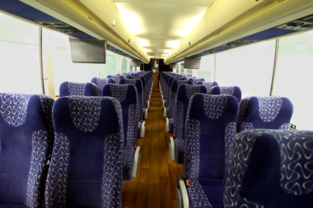 Interior of a charter bus cabin with rows of blue patterned seats, a center aisle, wood-look flooring, and overhead monitors