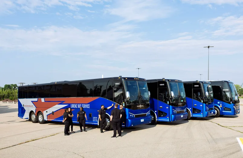Group of Lorenz Bus Service drivers in black uniforms gathered in front of a lineup of blue motorcoaches outdoors