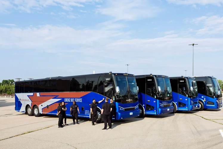 Four Lorenz Bus Service drivers in black uniforms standing in front of a mixed fleet of motorcoaches and minibuses lined up outdoors