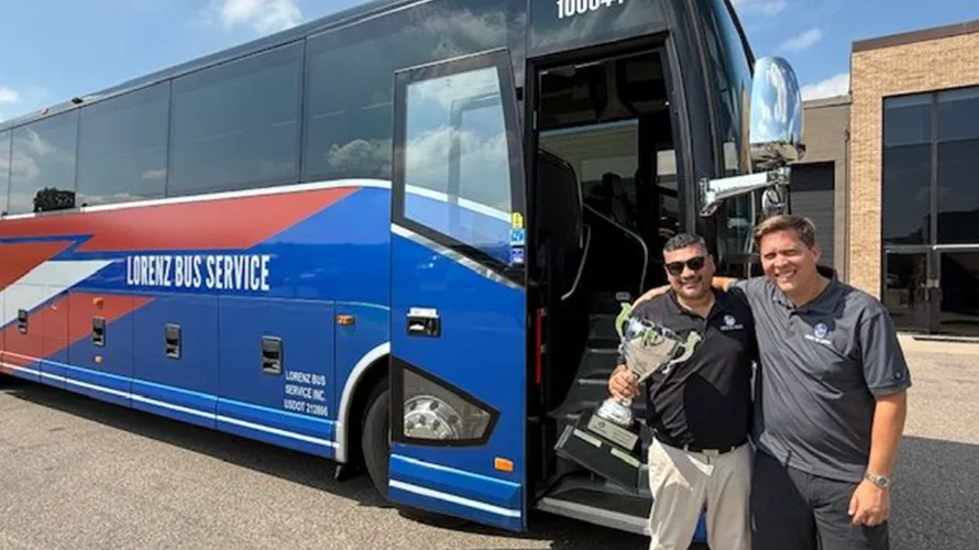 Two Lorenz Bus Service staff members holding a large trophy in front of a branded motorcoach outdoors