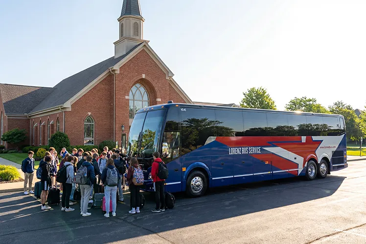 Students with backpacks and luggage boarding a Lorenz Bus Service coach outside a brick church building