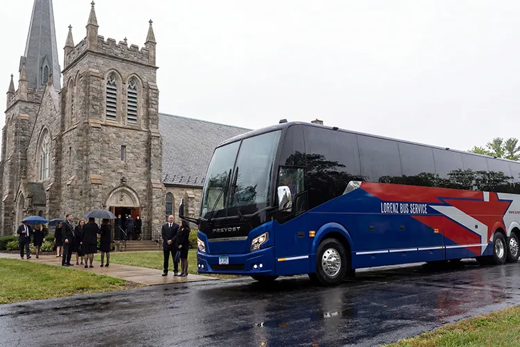 Lorenz Bus Service Prevost coach parked outside a stone church, with formally dressed guests gathering nearby on a rainy day