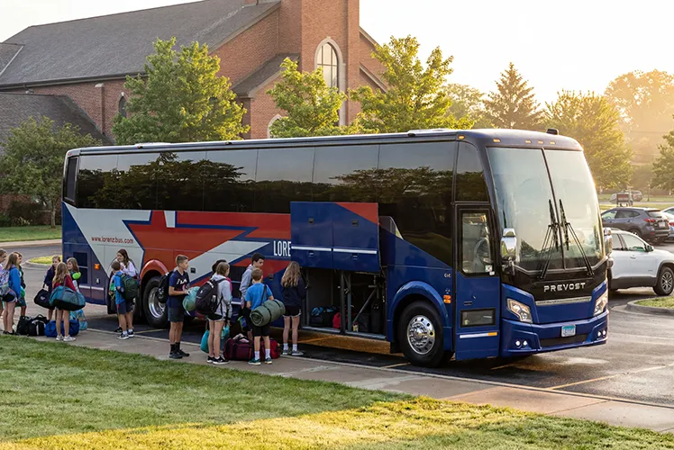 Students with backpacks and luggage boarding a Lorenz charter bus in a parking lot at sunrise