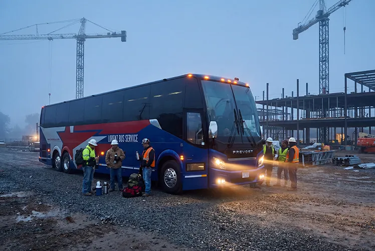 Lorenz Bus Service motorcoach parked at an active construction site at dawn, with crew members in hard hats and safety vests gathered alongside