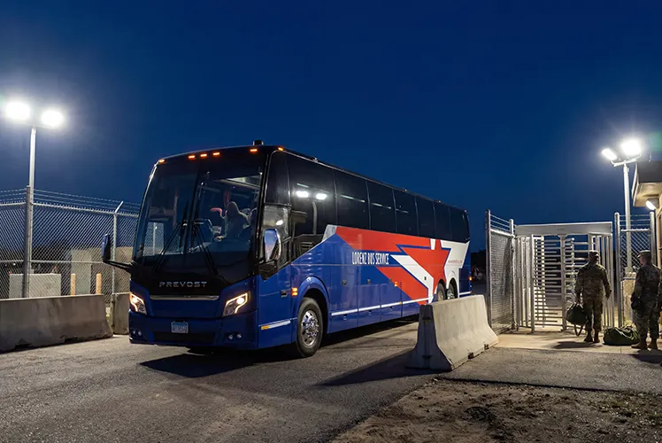Lorenz Bus Service Prevost coach entering a secured military base gate at night with personnel standing nearby