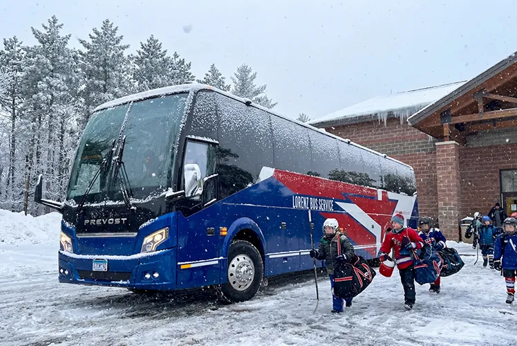 Youth hockey players in full gear boarding a Lorenz Bus Service Prevost coach in snowy winter conditions outside a rink