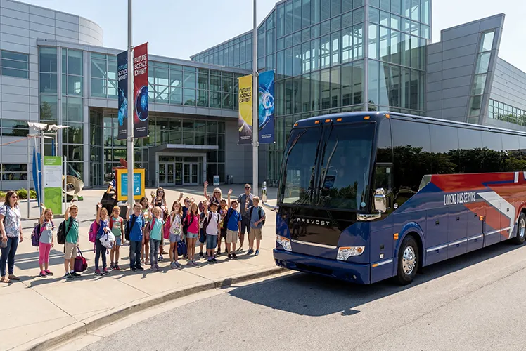 A group of elementary students and chaperones wave beside a Lorenz Bus Service coach outside a science museum on a field trip