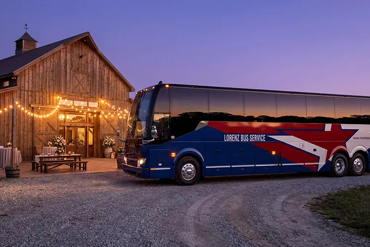 Lorenz Bus Service motorcoach parked outside a rustic barn wedding venue at dusk with string lights glowing at the entrance