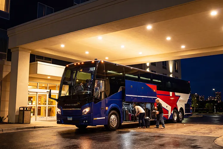 Prevost charter bus parked under a hotel canopy at night while passengers load luggage into the undercarriage storage