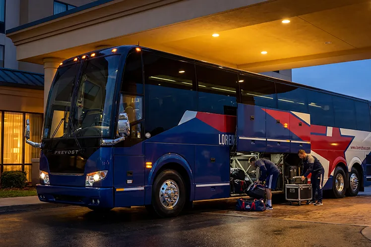 Lorenz Bus Service Prevost charter coach parked at a hotel while crew loads equipment into undercarriage storage at dusk