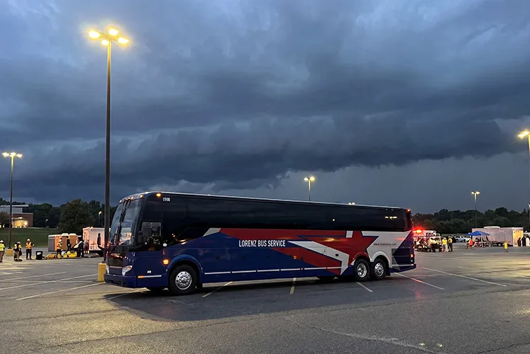 Lorenz Bus Service motorcoach staged in a parking lot at dusk beneath stormy skies, with event staff and equipment visible in the background