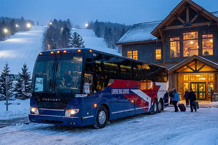 Lorenz Bus Service motorcoach parked at a ski resort lodge entrance in winter, with passengers unloading gear in the snow