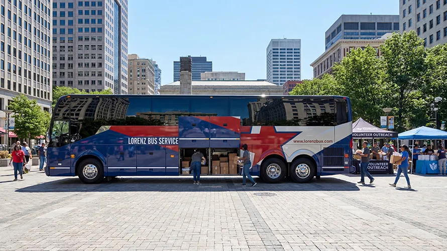 Lorenz Bus Service coach parked in a downtown plaza during a volunteer outreach event, with boxes being unloaded from the undercarriage storage