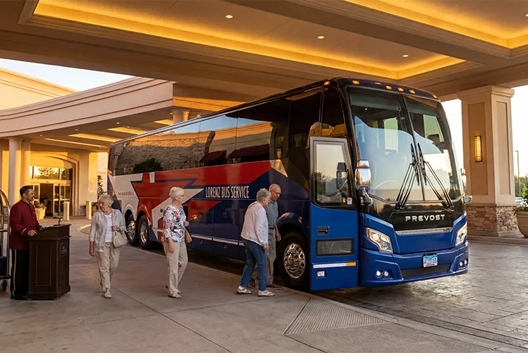 Passengers boarding a Lorenz Bus Service Prevost motorcoach under a hotel porte-cochère at dusk