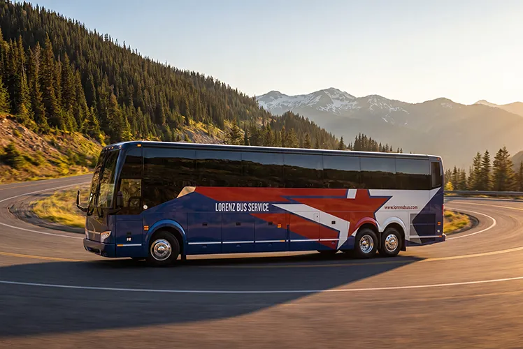 Lorenz Bus Service motorcoach traveling a mountain highway at sunset, with forested peaks in the background
