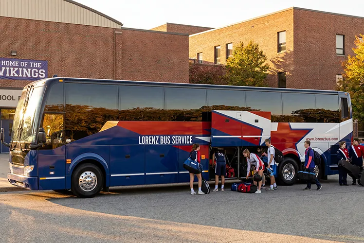 Athletes loading gear into the undercarriage storage of a Lorenz Bus Service coach outside a school gymnasium
