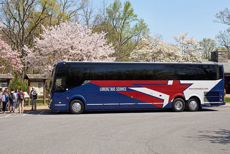 Lorenz Bus Service motorcoach parked at an outdoor destination as a group of passengers boards, with cherry blossoms in the background