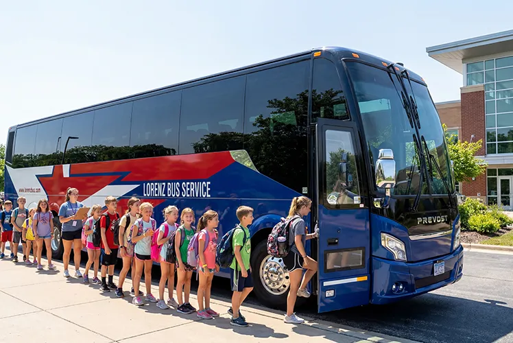 Students with backpacks boarding a Lorenz Bus Service Prevost motorcoach outside a school building on a sunny day