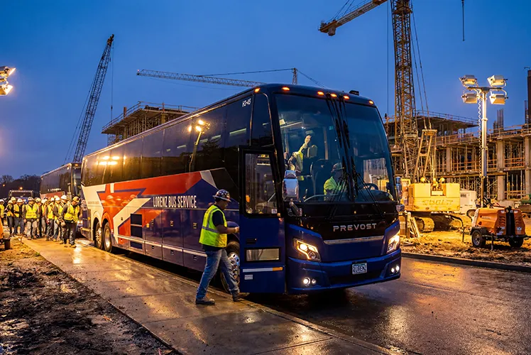 Lorenz Bus Service motorcoach at an active construction site at dusk, with crew workers in high-visibility vests boarding nearby