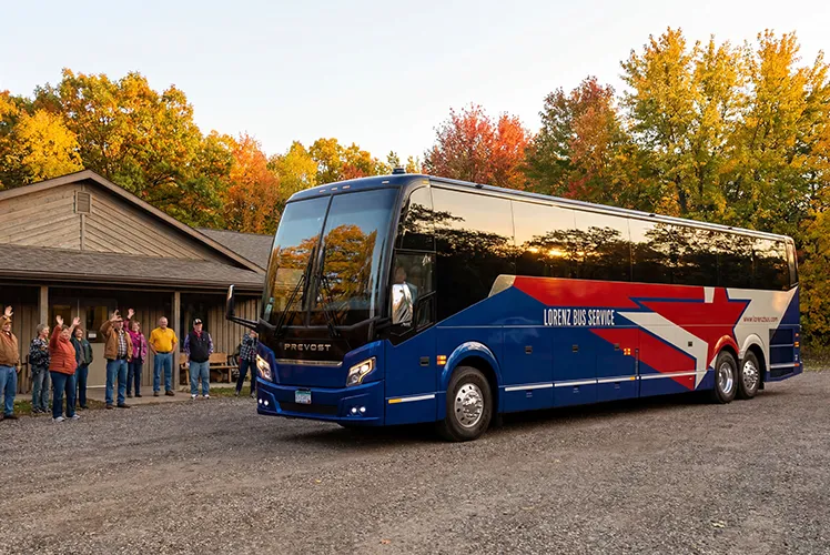 Lorenz Bus Service Prevost motorcoach parked outside a lodge as a group of passengers waves goodbye, surrounded by fall foliage