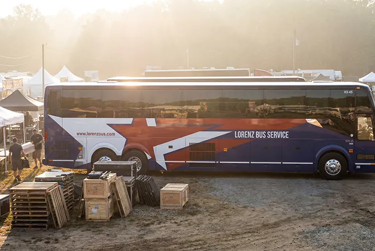 Lorenz Bus Service coach parked at an outdoor event staging area surrounded by equipment crates and vendor tents at sunrise
