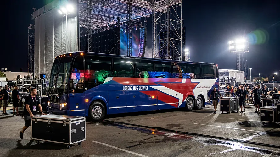 Lorenz Bus Service coach parked backstage at a concert venue at night, with stage rigging and crew members nearby