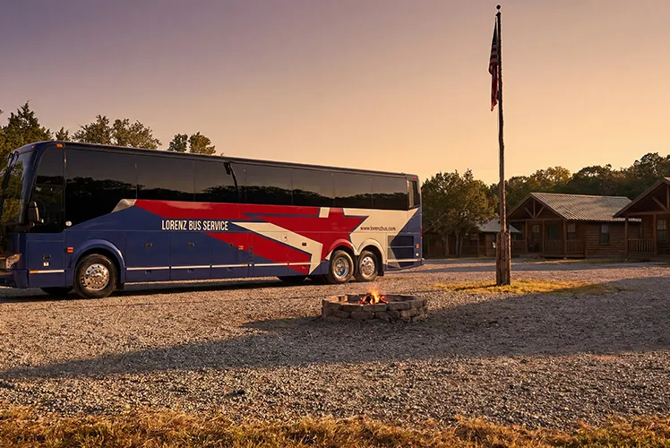 Lorenz Bus Service charter coach parked at a rustic camp setting with a campfire, flagpole, and log cabin at sunset