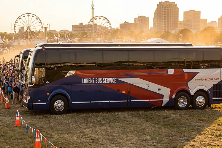 Lorenz Bus Service coach parked at an outdoor festival grounds at sunset, with a crowd boarding and a city skyline in the background