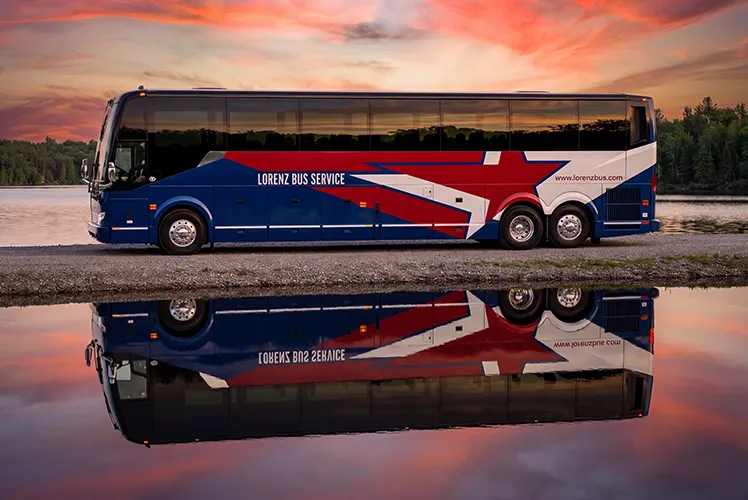 Lorenz Bus Service full-size motorcoach parked at sunset, its red, white, and blue wrap reflected in still water below