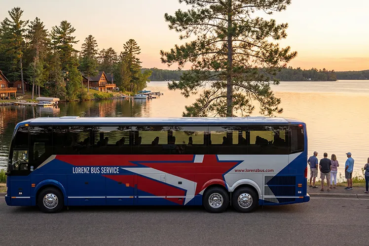 Lorenz Bus Service motorcoach parked lakeside at sunset with passengers boarding near pine trees and cabin-lined shore