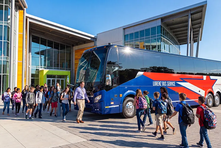 Students with backpacks boarding a Lorenz Bus Service motorcoach outside a school building with an adult chaperone nearby