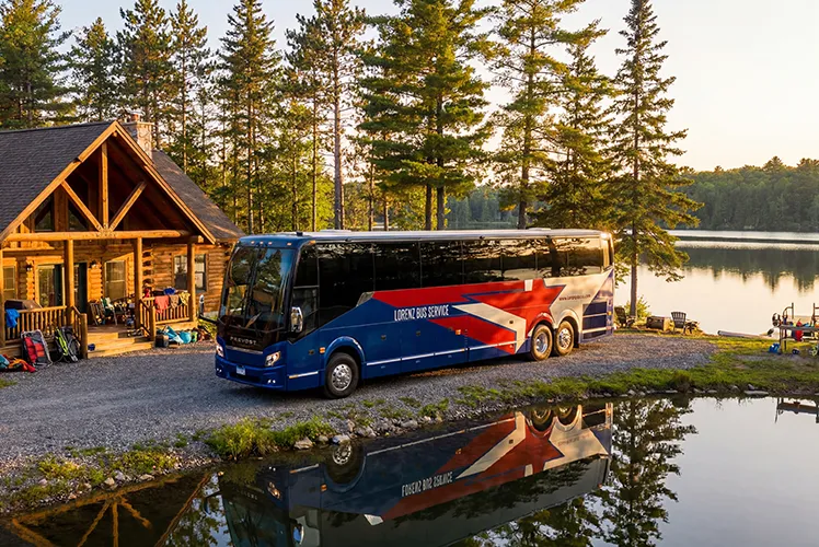 Lorenz Bus Service motorcoach parked at a lakeside log cabin surrounded by pine trees at sunset
