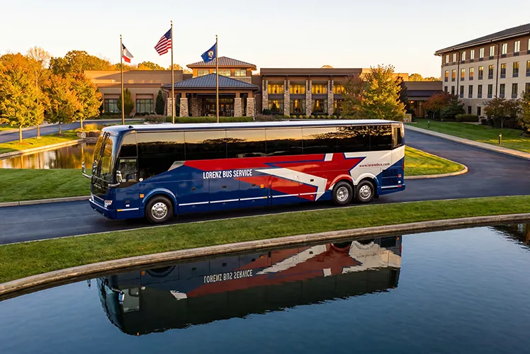 Lorenz Bus Service motorcoach parked in front of a resort-style venue, reflected in a still pond at golden hour