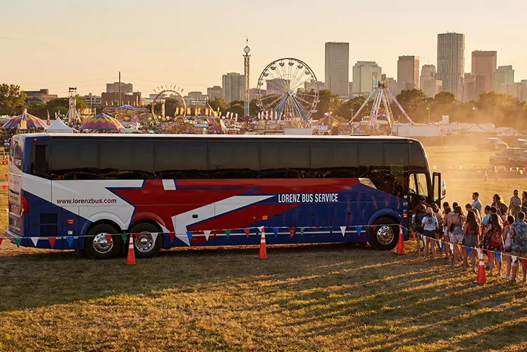 Lorenz Bus Service charter coach parked at an outdoor festival grounds at sunset, with attendees boarding and a city skyline in the background