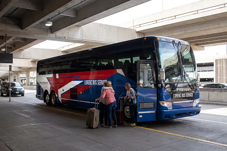 Two passengers with luggage boarding a Lorenz Bus Service Prevost coach at an airport pickup area