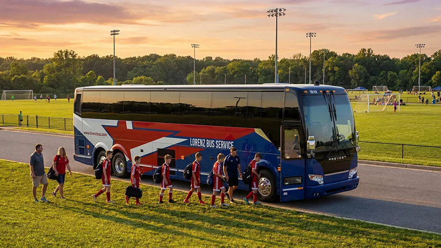 Youth soccer players in red uniforms boarding a Lorenz Bus Service Prevost coach at a soccer complex at sunset