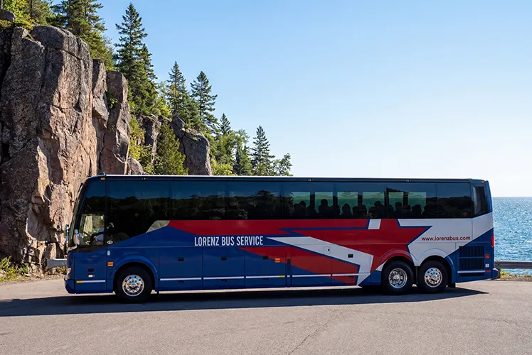 Lorenz Bus Service full-size motorcoach parked along a scenic rocky shoreline with pine trees and blue water